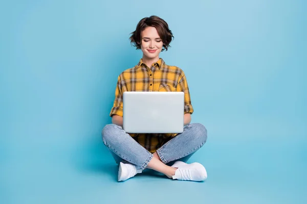 Photo of charming freelancer lady sit floor wpisując netbook nosić koszulę w kratę dżinsy buty odizolowany niebieski kolor tło — Zdjęcie stockowe