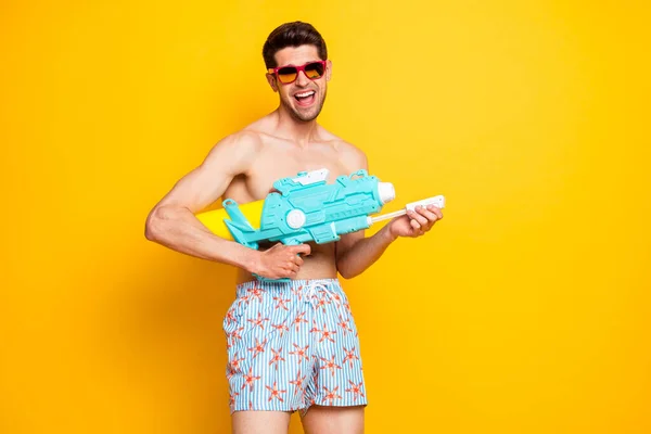 Foto von niedlichen lustigen jungen Mann nackten Oberkörper tragen dunkle Brille Schießen Wasserpistole lächelt isoliert gelbe Farbe Hintergrund — Stockfoto