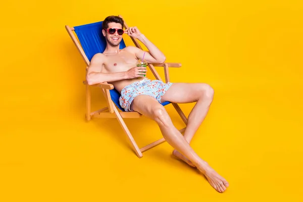 Foto de lindo chico dulce sin camisa vestida gafas oscuras tumbado silla de salón beber cóctel aislado color amarillo fondo — Foto de Stock
