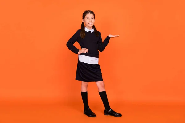 Foto av bedårande skolflicka hand presentera tomt utrymme bära enhetlig isolerad orange färg bakgrund — Stockfoto