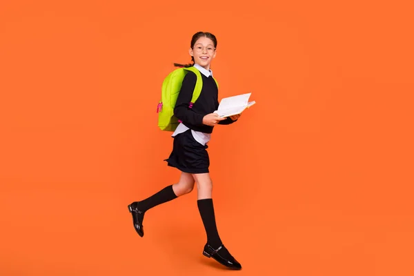 Ganzkörperfoto von fröhlichen brünetten Zöpfen haarige Mädchen springen nach oben halten Buch tragen Tasche isoliert auf orangefarbenem Hintergrund — Stockfoto