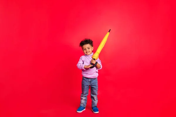 Повнометражний вигляд на тіло привабливого веселого хлопчика, що тримає парасольковий дитячий садок, ізольований на яскраво-червоному кольоровому фоні — стокове фото