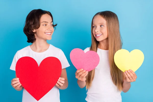 Foto de duas crianças românticas segurar corações de papel olhar uns aos outros desgaste branco t-shirt isolado azul cor de fundo — Fotografia de Stock