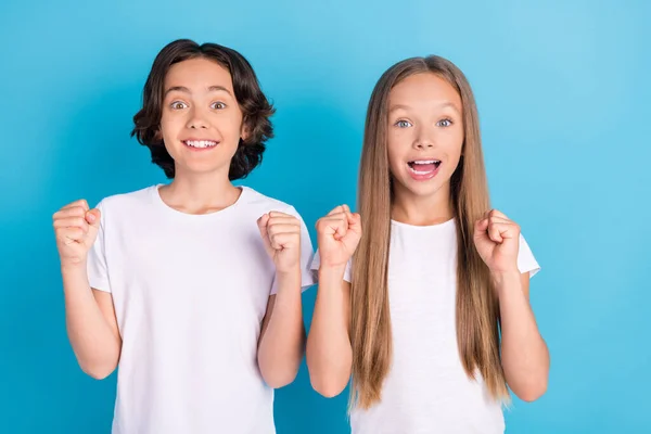 Foto de dos niños asombrados celebran victoria levantan los puños usan camiseta blanca aislado fondo de color azul —  Fotos de Stock