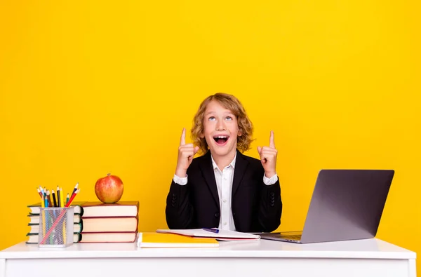 Foto van opgewonden kind zitten bureau geven vinger lege ruimte op te zoeken dragen uniform geïsoleerde gele kleur achtergrond — Stockfoto