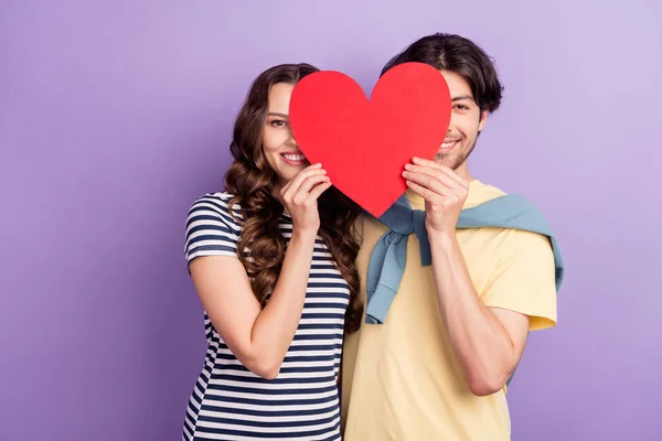 Photo of adorable shy couple wear casual clothes smiling holding large paper heart cover eyes isolated violet color background — Stock Photo, Image