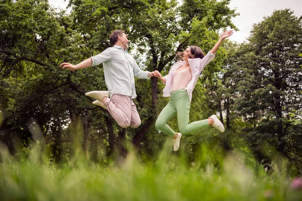Фотография блестящей беззаботной пары носить повседневную одежду улыбаясь держа руки прыгая высоко на открытом воздухе городской парк — стоковое фото