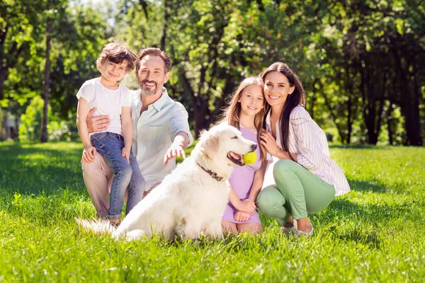 Foto de familia amable dulce vestido traje casual sentado hierba verde lanzando bola de perro sonriendo fuera de la calle urbana de la ciudad — Foto de Stock