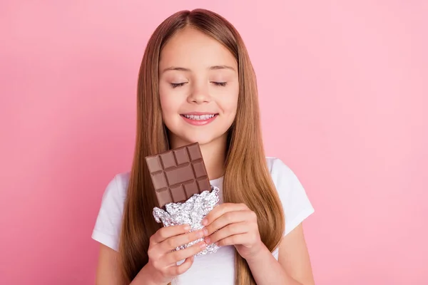 Sarışın, küçük bir kızın çikolata yerken çekilmiş fotoğrafı. Kapalı gözler. Pembe arka planda izole edilmiş beyaz tişört giyiyor. — Stok fotoğraf
