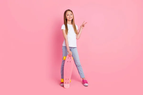 Full storlek foto av cool blond liten flicka hålla skridsko show v-tecken bära vit t-shirt jeans isolerad på rosa färg bakgrund — Stockfoto