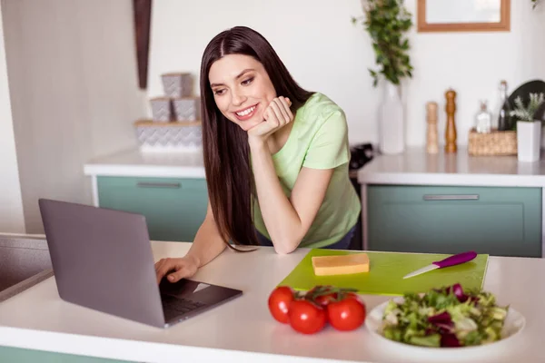 Φωτογραφία από αρκετά αξιολάτρευτο νεαρή γυναίκα ντυμένος πράσινο t-shirt μαγείρεμα πρωινό δακτυλογράφηση σύγχρονο gadget σε εσωτερικούς χώρους σπίτι δωμάτιο — Φωτογραφία Αρχείου