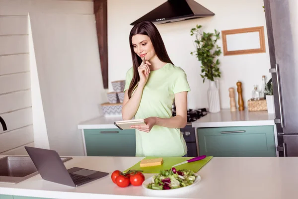 Φωτογραφία της σκέψης μακρύ χτένισμα νεαρή κοπέλα ματιά φορητό υπολογιστή γράψει μάγειρας φορούν πράσινο t-shirt στην κουζίνα και μόνο στο σπίτι — Φωτογραφία Αρχείου