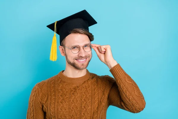 Foto av självsäker söt kille klädd stickad tröja akademisk huvudbonad arm glasögon ler isolerad blå färg bakgrund — Stockfoto