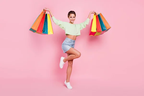 Full längd foto av glad söt ung kvinna höja händerna hålla väskor shopper isolerad på rosa färg bakgrund — Stockfoto