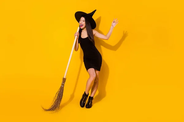 Pełne ciało zdjęcie śpiewać czarownica młoda dama z miotła nosić czarny sukienka czapka buty izolowane na żółty kolor tło — Zdjęcie stockowe