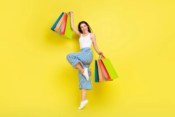 Volledige grootte foto van onder de indruk brunette millennial dame springen hold tassen dragen top broek sneakers geïsoleerd op geel kleur achtergrond — Stockfoto