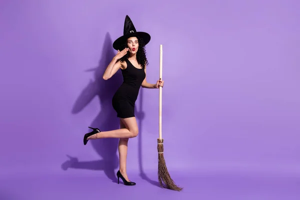 全长照片，漂亮的黑发女士站在与扫帚穿黑色连衣裙鞋帽隔离在紫丁香背景印象深刻 — 图库照片