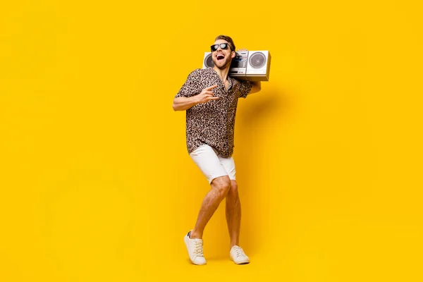 Teljes hosszúságú fotó jóképű hűvös úriember öltözött nyomtatott ing szemüveg mutatja kemény rock jel boombox izolált sárga színű háttér — Stock Fotó