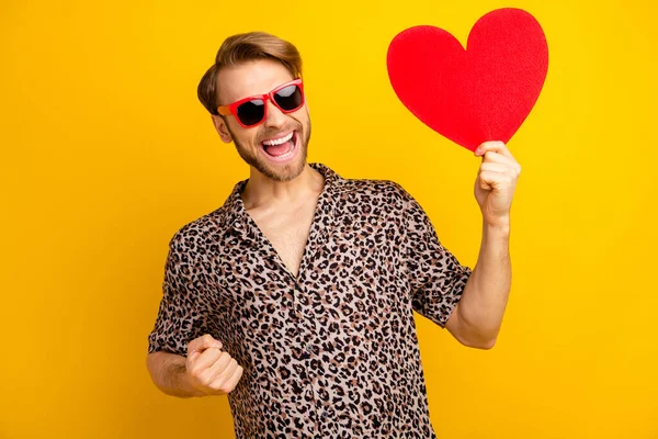 Photo of impressed sweet guy wear leopard outfit arm dark eyewear rising fists holding red heart isolated yellow color background — Stock Photo, Image