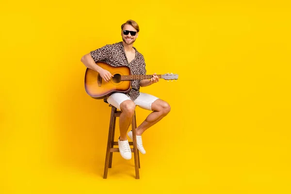 Foto de bastante guapo joven llevar traje de leopardo gafas oscuras silla de bar sentado tocando la guitarra aislado color amarillo fondo —  Fotos de Stock