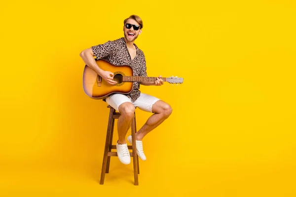 Photo of sweet funny young gentleman dressed print shirt dark glasses sitting bar krzesło gra na gitarze śpiew odizolowany żółty kolor tła — Zdjęcie stockowe