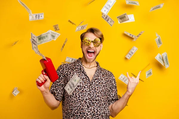 Foto de guapo rudo caballero vestido camisa gafas oscuras tiro pistola de dinero mostrando señal de roca aislado color amarillo fondo — Foto de Stock