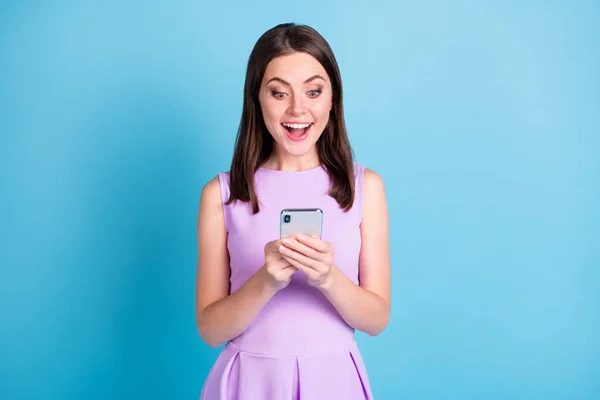 Фотографія здивованої позитивної молодої леді в фіолетовому вбранні тримає новинку смартфона ізольовано на синьому кольоровому фоні — стокове фото
