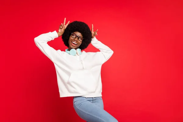 Фото милой кудрявой причёски Millennial леди показать V-знак держать наушники носить очки белой спортивной одежды изолированы на красном фоне — стоковое фото