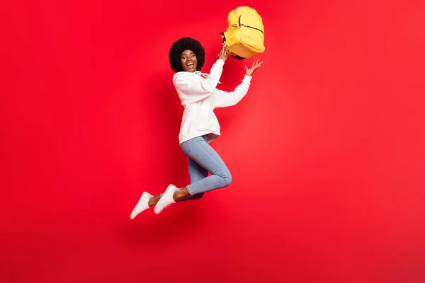 Полный профиль тела фото прохладной волнистой прической студентки прыжок с рюкзаком носить белые джинсы спортивной одежды изолированы на красном фоне — стоковое фото