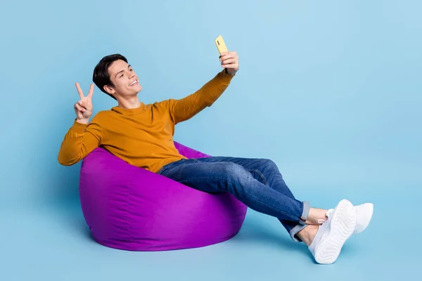 Foto de longitud completa de la divertida morena millennial chico sentarse en el bolso hacer selfie v-signo de desgaste pantalones vaqueros aislados en el fondo de color azul —  Fotos de Stock