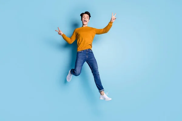Полноразмерная фотография сумасшедшего брюнета-миллениала, который прыгает, показывает, что он носит свитер в джинсах на голубом цветном фоне — стоковое фото