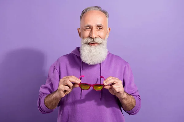 Portrait of attractive cheerful grey-haired man holding specs wearing season clothes isolated over purple violet color background — Stock Photo, Image