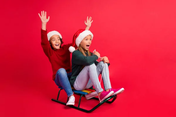 Foto de colegiales dulces emocionados vestidos suéteres montando trineos levantando las manos buscando espacio vacío aislado color rojo fondo — Foto de Stock