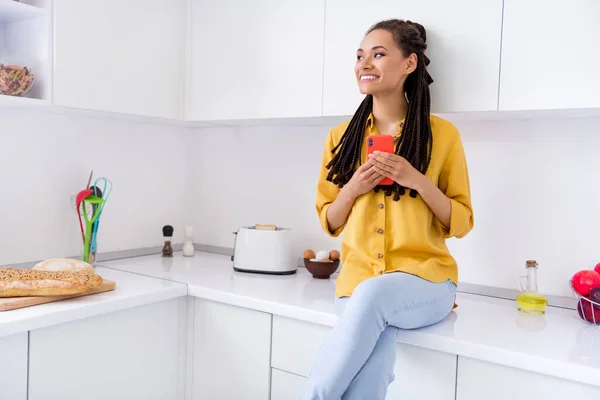 Foto de mulher de pele escura sonhadora engraçada vestida camisa amarela mesa de estar segurando gadget moderno dentro de casa casa quarto — Fotografia de Stock