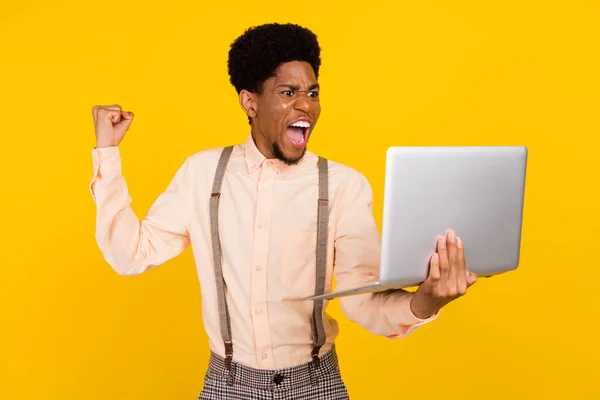 Foto de fã alegre cara segurar netbook grito levantar punho desgaste suspensórios camisa isolado cor amarela fundo — Fotografia de Stock