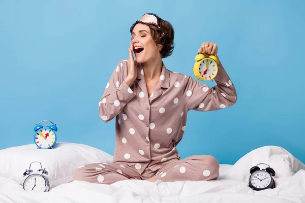 Foto di giovane felice felice sorridente ragazza funky sbadigliare sedersi sul letto circondato da orologi isolati su sfondo di colore blu — Foto Stock