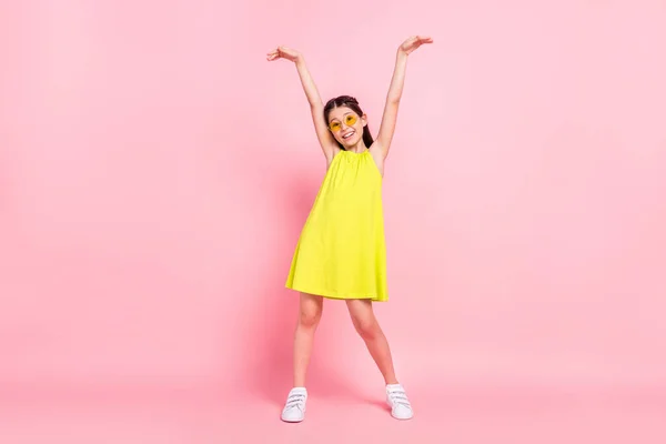 Full length φωτογραφία του γλυκού λαμπερό κορίτσι σχολείο φορούν κίτρινο φόρεμα γυαλιά χαμογελαστό χορό αύξηση χέρια απομονωμένο ροζ χρώμα φόντο — Φωτογραφία Αρχείου