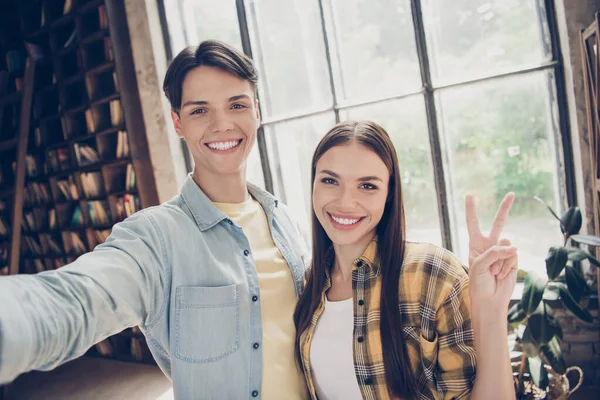 Autorretrato de dos personas alegres y atractivas que visitan la biblioteca mostrando el signo v en el interior industrial loft en el espacio abierto — Foto de Stock