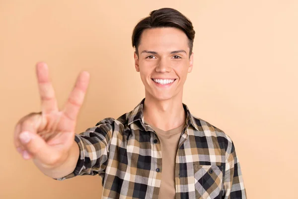 Foto di ragazzo divertente millenario mostra v-segno indossare camicia a scacchi isolato su sfondo di colore beige — Foto Stock