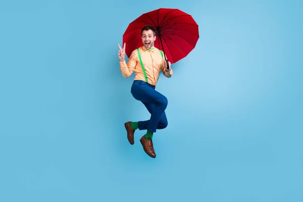 Ganzkörperfoto von flippigen brünetten Mann springen mit Sonnenschirm zeigen V-Zeichen tragen Hemd Hose Socken Schuhe isoliert auf blauem Hintergrund — Stockfoto