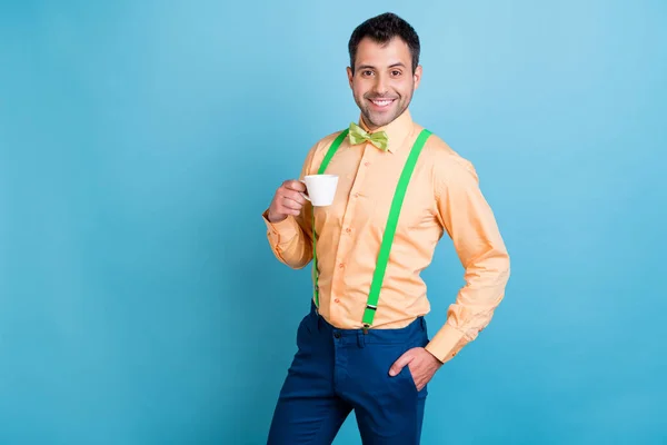 Foto de chico alegre mantenga taza bebida café desgaste tirantes verdes camisa lazo aislado azul color fondo —  Fotos de Stock