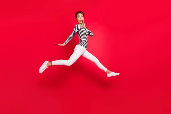 Foto de brillante mujer joven impresionado vestido a rayas brazo mejilla saltar hign aislado color rojo fondo —  Fotos de Stock