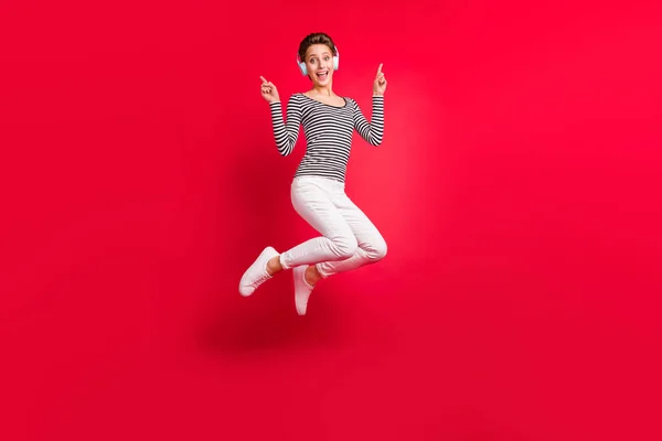 Foto de mujer bastante dulce vestida a rayas auriculares de traje saltando señalando los dedos espacio vacío aislado color rojo fondo —  Fotos de Stock