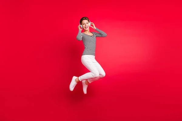 Foto de brillante divertida joven dama usar ropa a rayas auriculares sonriendo saltando alto aislado color rojo fondo —  Fotos de Stock