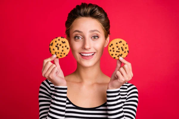 Foto de engraçado encantador jovem senhora usar roupas listradas segurando dois biscoitos saborosos sorrindo isolado fundo cor vermelha — Fotografia de Stock