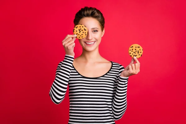 Foto de menina engraçada bonito usar roupas listradas segurando dois biscoitos saborosos cobrir o olho sorrindo isolado fundo cor vermelha — Fotografia de Stock