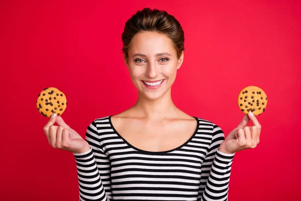 Fénykép imádnivaló aranyos fiatal hölgy viselni csíkos ruhát gazdaság két ízletes cookie mosolygós elszigetelt piros színű háttér — Stock Fotó