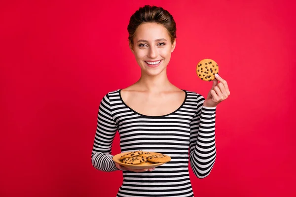 Foto de excitado encantador jovem mulher vestida roupa listrada segurando casa fez cookies isolado fundo cor vermelha — Fotografia de Stock