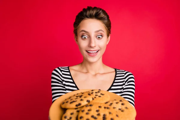 Foto di dolce eccitato giovane donna vestito vestito a righe tenendo casa fatto biscotti grandi occhi enormi isolato colore rosso sfondo — Foto Stock