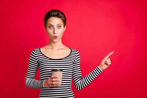Φωτογραφία από γλυκό εντυπωσιασμένος γυναίκα ντυμένος ριγέ στολή κρατώντας φλιτζάνι καφέ δείχνοντας κενό χώρο δάχτυλο απομονωμένο φόντο κόκκινο χρώμα — Φωτογραφία Αρχείου